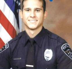 Portrait of a uniformed police officer in a formal studio photograph, standing in front of an American flag and wearing a dark police uniform with badge and tie.