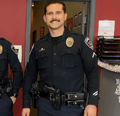 Police officer in uniform smiling while standing indoors near an office doorway. The officer wears a badge, duty belt, and department patches.