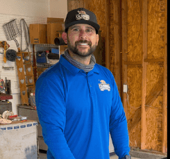 SBVC alumnus wearing a blue company polo shirt and branded cap stands inside a construction site, smiling in front of unfinished wood framing and building materials.