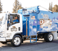 Blue service truck with company branding parked outdoors. A driver wearing a company uniform leans out of the driver's side window and smiles toward the camera.