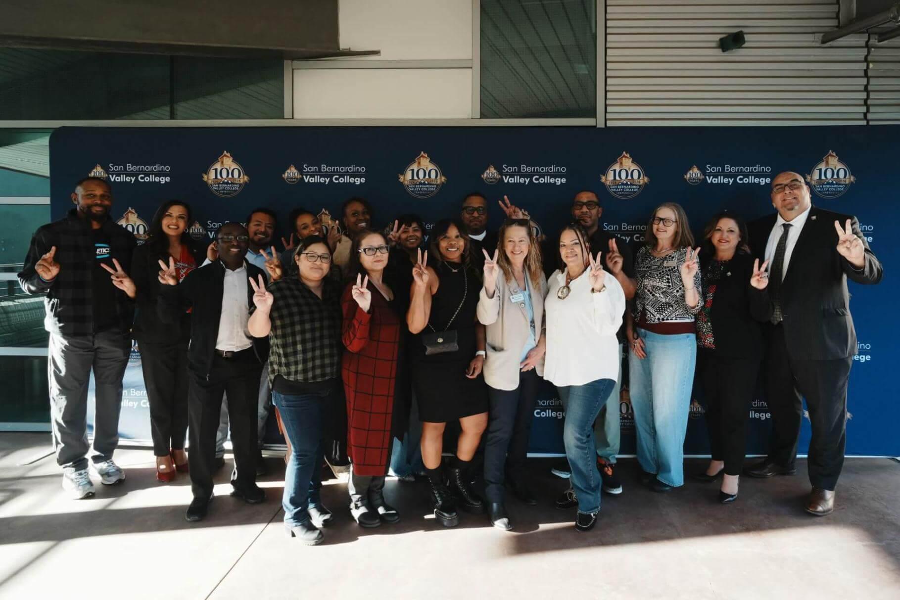 Approximately 17 San Bernardino Valley College faculty and staff members pose together in front of a blue Centennial step-and-repeat banner displaying the SBVC logo and 100-year anniversary emblem. The group stands outside a modern campus building, smiling and holding up peace signs in celebration. Natural sunlight casts shadows across the foreground while the Centennial branding remains prominently visible behind them.