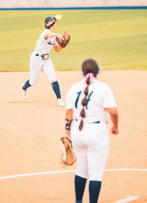 An SBVC softball player in a striped uniform throws a softball across the infield while a teammate waits in the foreground. The action takes place on a sunny day, with the dirt infield and green outfield visible in the background.