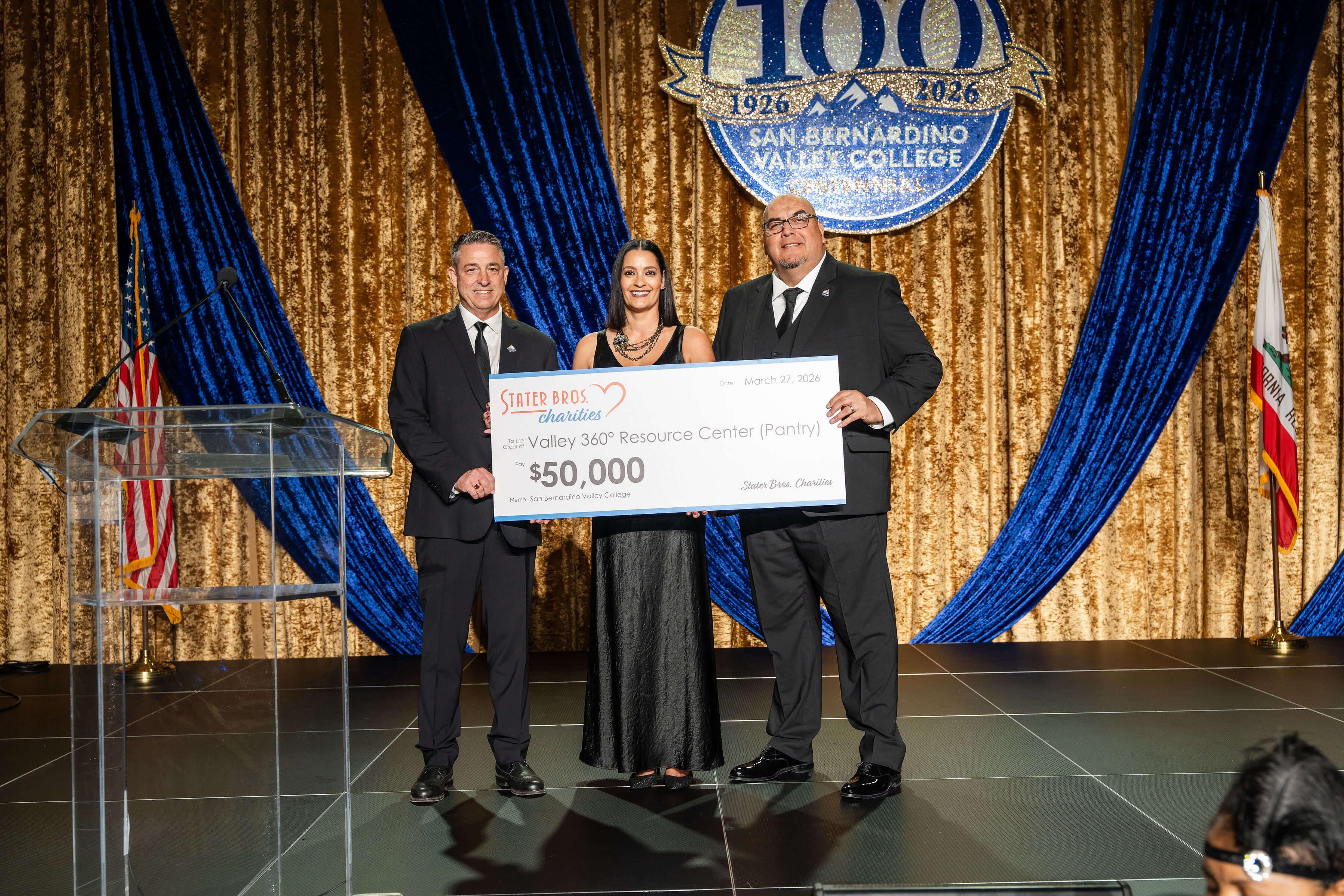 During the SBVC Centennial Gala, three presenters stand on stage holding a ceremonial $50,000 donation check from Stater Bros. Charities for the Valley 360° Resource Center Pantry, framed by blue drapery and Centennial branding.