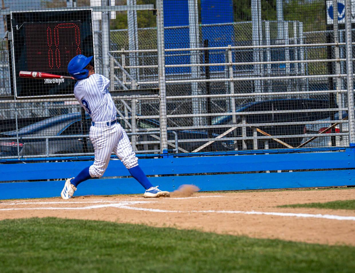 An SBVC Wolverines baseball player in a blue-and-white striped uniform follows through on a swing during a game as dirt rises from the batter’s box and stadium seating fills the background.