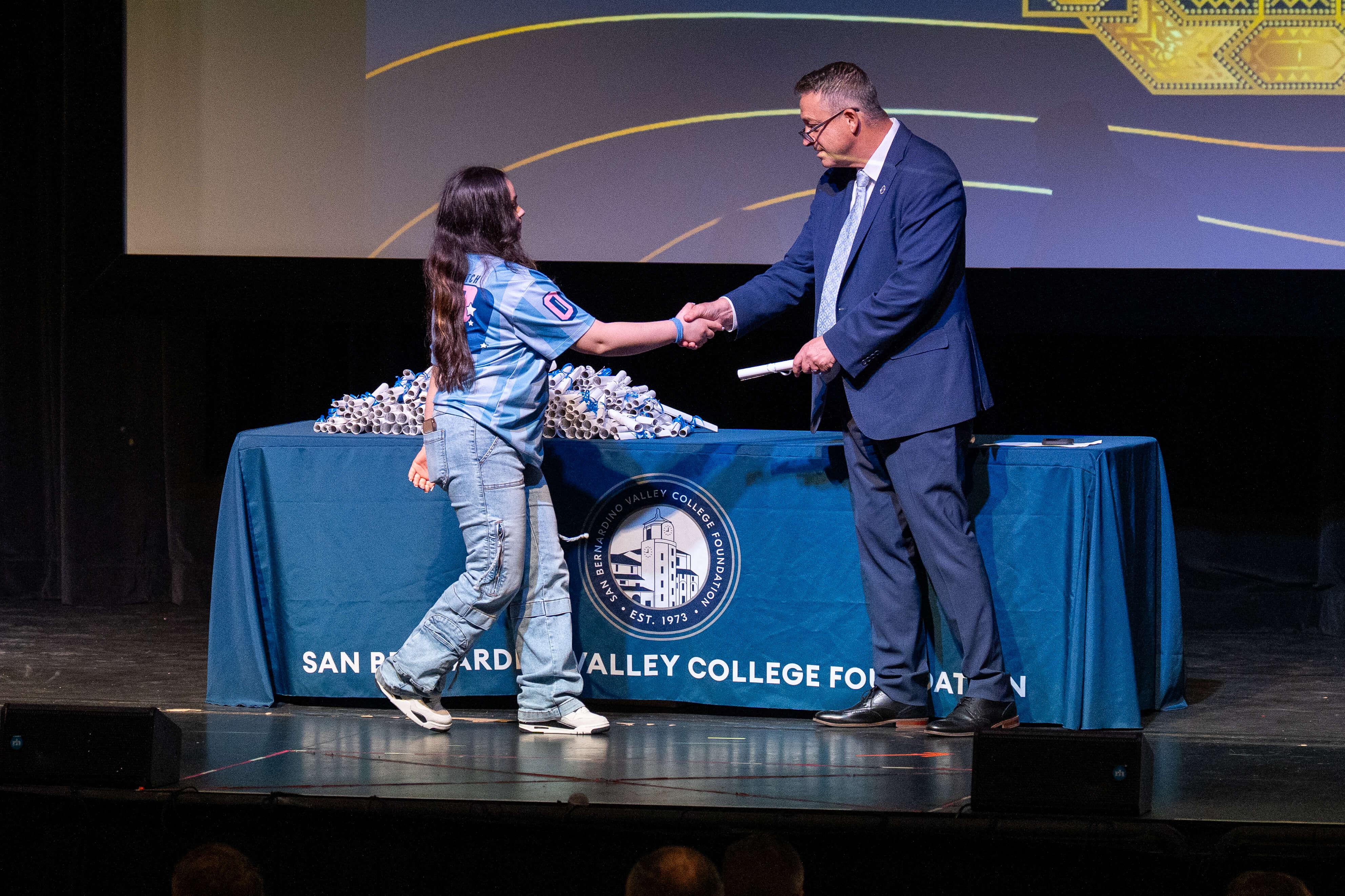 An SBVC Foundation scholarship recipient shakes hands with a college representative on stage while receiving recognition. Scholarship certificates are arranged on a table behind them during the annual awards ceremony.